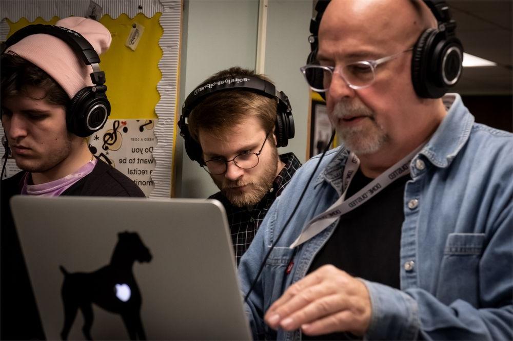 A professor with headphones works on a computer while two students also wearing headphones work alongside him.